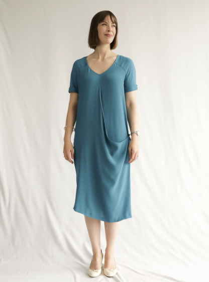 Woman wearing the Maeve Woven Dress sewing pattern from Style Arc on The Fold Line. A Dress pattern made in viscose, silk or crepe fabrics, featuring a slip-on style, V-neck, front and back yokes extending into sleeves, turn-up cuffs, inverted back pleat, front drape feature with loop for buttoning to side seam button, and midi length.