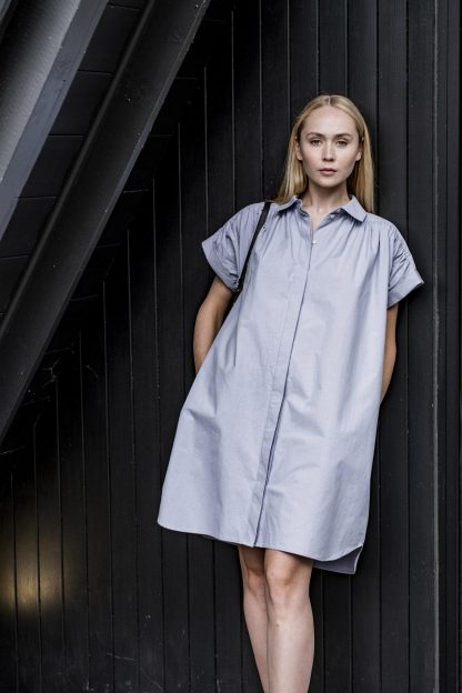 Woman wearing the Augusta Dress sewing pattern from Fibre Mood on The Fold Line. A shirtdress pattern made in chambray, linen, poplin, cotton voile, crepon, crepe, supple twill, double gauze, seersucker, or washed silk fabrics, featuring rolled short sleeves, collar with collar band, concealed front button fastening, side seam pockets and gathered front and back yoke.