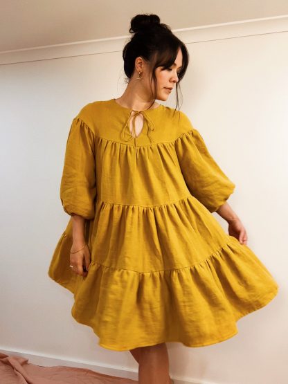 Woman wearing the Hazel Dress sewing pattern from Veronica Tucker on The Fold Line. A dress pattern made in medium weight opaque fabrics, featuring a tie neck yoke, balloon sleeves, three voluminous tiers, pockets, and knee length hem.