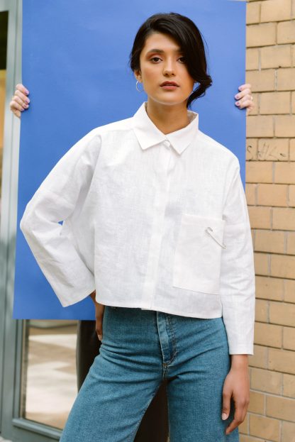 Woman wearing the Noella Blouse sewing pattern from JULIANA MARTEJEVS on The Fold Line. A shirt pattern made in cotton poplin fabrics, featuring an oversized, cropped silhouette, long sleeves, single front pocket, stand collar, back yoke with inverted pleat and button front closure.