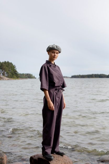 Woman wearing the Judith Jumpsuit sewing pattern from TAUKO on The Fold Line. A jumpsuit pattern made in cotton, cotton/polyester blend, linen, hemp, or light denim fabrics, featuring a pointed collar, snap button placket, side pockets, self-fabric belt, elbow length sleeves, back yoke, straight legs and front and back waist darts.