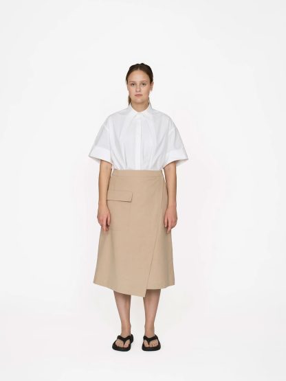 Woman wearing the Asymmetric Midi Skirt sewing pattern from The Assembly Line on The Fold Line. A wrap skirt pattern made in cotton, denim, wool and faux leather fabrics, featuring a midi length, asymmetric silhouette, back darts, curved waistband with snap button closure, and front patch pocket with flap.