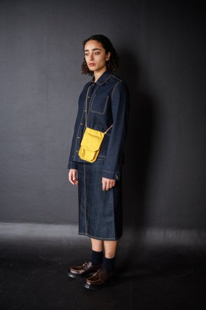 Woman wearing the Francli Pocket Bag sewing pattern from Merchant & Mills on The Fold Line. A bag pattern made in oilskin, dry oilskin, cotton canvas or drill, and denim fabrics, featuring an inner pocket, magnetic snap fastening, and sliding knot strap.