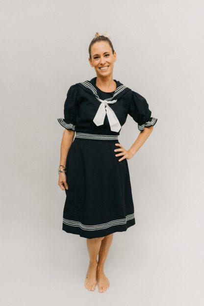 Woman wearing the 253 Vintage Bathing Costume sewing pattern from Folkwear on The Fold Line. A bathing suit pattern made in medium weight cottons, linens and blends fabrics, featuring a gored skirt with deep back pleats and buttoned waistband. Knee length romper has front button closure, short puff sleeves, sailor collar and tie.