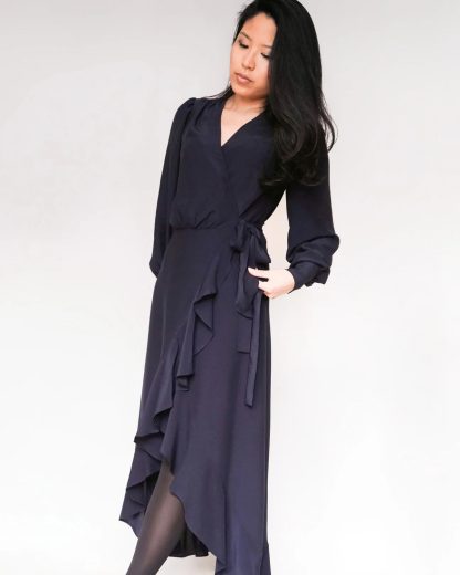 Woman wearing the Joline Dress sewing pattern from Bara Studio on The Fold Line. A wrap dress pattern made in viscose, cotton, linen, or lyocell fabrics, featuring a deep V-neck, tiered high-low hem, full length sleeve, gathered cuff with button closure, self-fabric waist tie and.