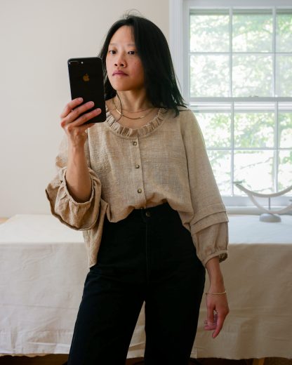 Woman wearing the Nepheline Blouse sewing pattern from Vivian Shao Chen on The Fold Line. A blouse pattern made in cotton lawn, batiste, voile, silk crepe de chine, silk georgette, poplin, or linen fabric, featuring a loose oversized fit, A-line silhouette, 3/4 length sleeves with tucks, and a scooped neckline with a ruffle.