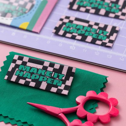 Photo of the 'Make It Happen' woven labels from Little Rosy Cheeks on The Fold Line. The pack includes 6 woven labels with green text on a black and pink chequered background, ready to be sewn into your handmade clothes.